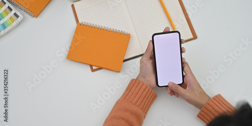 Top view image of creative woman hands holding/using a white blank screen smartphone over white working desk that surrounded by notebook, note, color guide and pencil as background.