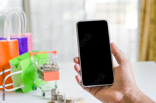 Hand holding mobile phone and In a small cart with paper bag and coin have on it online shopping located in home concept of detention at home to prevent the Covid19 virus.