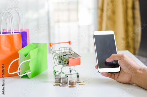 Hand holding mobile phone and In a small cart with paper bag and coin have on it online shopping located in home concept of detention at home to prevent the Covid19 virus.