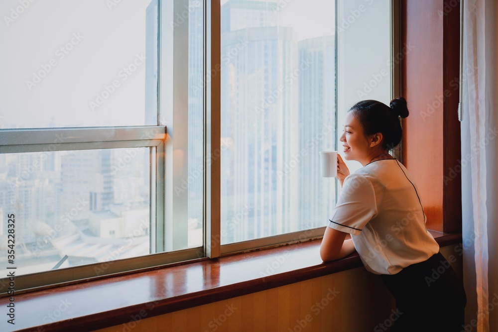 Asian woman holding coffee cup in bedroom looking out at window.Happy & smiling during the quarantine crisis of the covid19. Concept of social distancing at home in epidemic situation of corona virus. Asian woman holding coffee cup in bedroom looking out at window.Happy & smiling during the quarantine crisis of the covid19. Concept of social distancing at home in epidemic situation of corona virus.