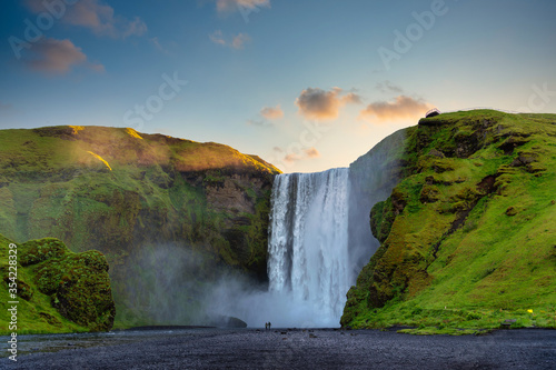 Panoramic view of the Skogafoss waterfall in Iceland. In the morning, the sunrise from behind a mountain with green grass, two male and female tourists are walking. This is a famous tourist attraction