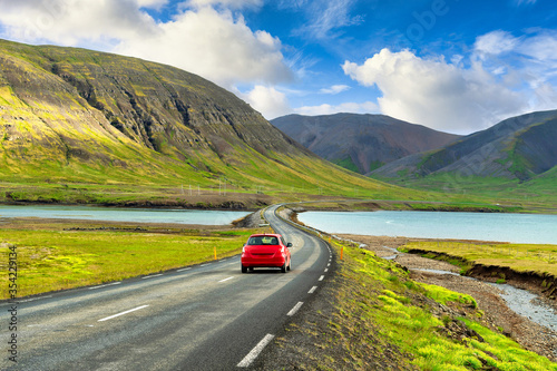 A red car runs on the highway in the Icelandic countryside. Amidst the great nature of mountains and lakes, in the summer there is grass all over In the concept road trip Holiday driving