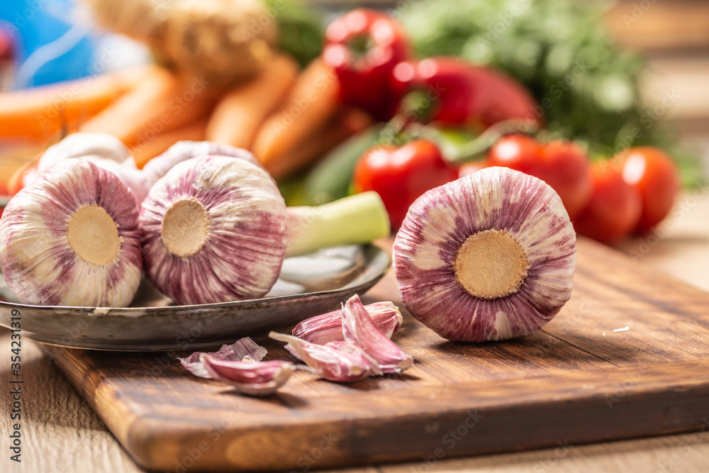 Five heads of garlic on a cutting board with various vegetables in the background