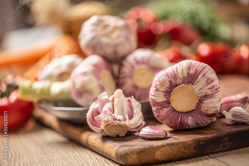 Five heads of garlic and one peeled garlic on a cutting board with various vegetables in the background