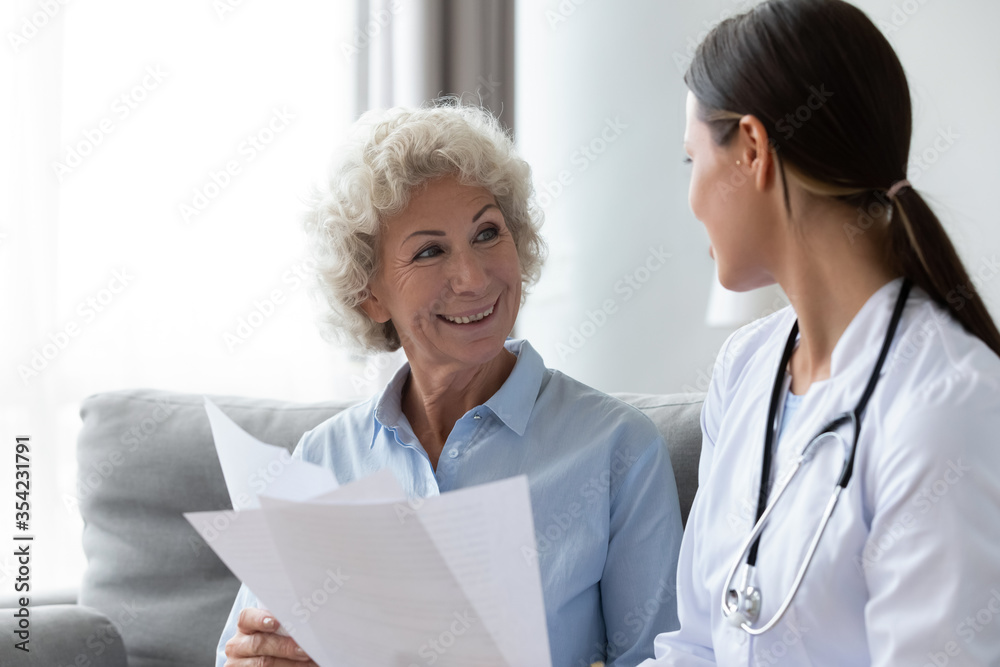Young nurse in white coat and stethoscope on shoulders after check up talking to elderly patient sitting on sofa tell about treatment plan, medication list, explain prescription and diagnosis concept - obrazy, fototapety, plakaty 