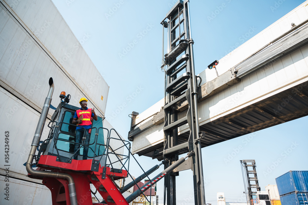 Black foreman worker working control the crane and forklift at Container cargo harbor to loading containers. African dock male staff for Logistics import export shipping concept.