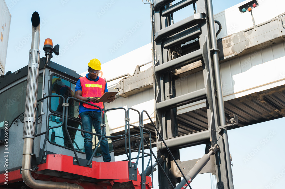Black foreman worker working control the crane and forklift at Container cargo harbor to loading containers. African dock male staff for Logistics import export shipping concept.