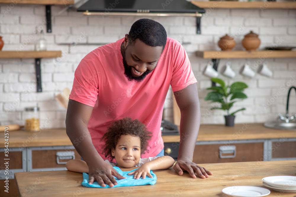 Fototapeta premium Tall dark-skinned man in a pink tshirt standing and wiping the table while his kid sitting
