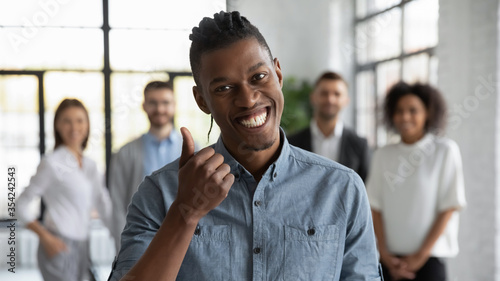 Head shot portrait smiling African American employee showing thumb up, standing with colleagues in modern office room, looking at camera, recommending corporate service, good career, human resources
