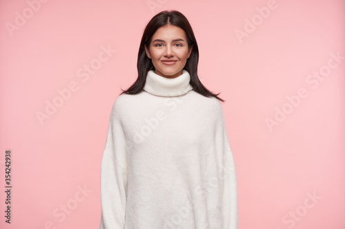Indoor shot of young charming brown haired woman dressed in white wollen poloneck looking positively at camera with pleasant smile, standing over pink background