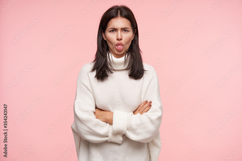 Offended young brown haired woman with casual hairstyle keeping her hands crossed and showing tongue to camera while standing against pink background Offended young brown haired woman with casual hairstyle keeping her hands crossed and showing tongue to camera while standing against pink background