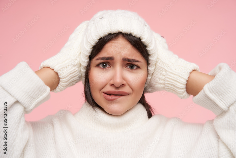 Confused young pretty brown haired woman dressed in white knitted clothes biting her underlip while looking worringly at camera, isolated over pink background