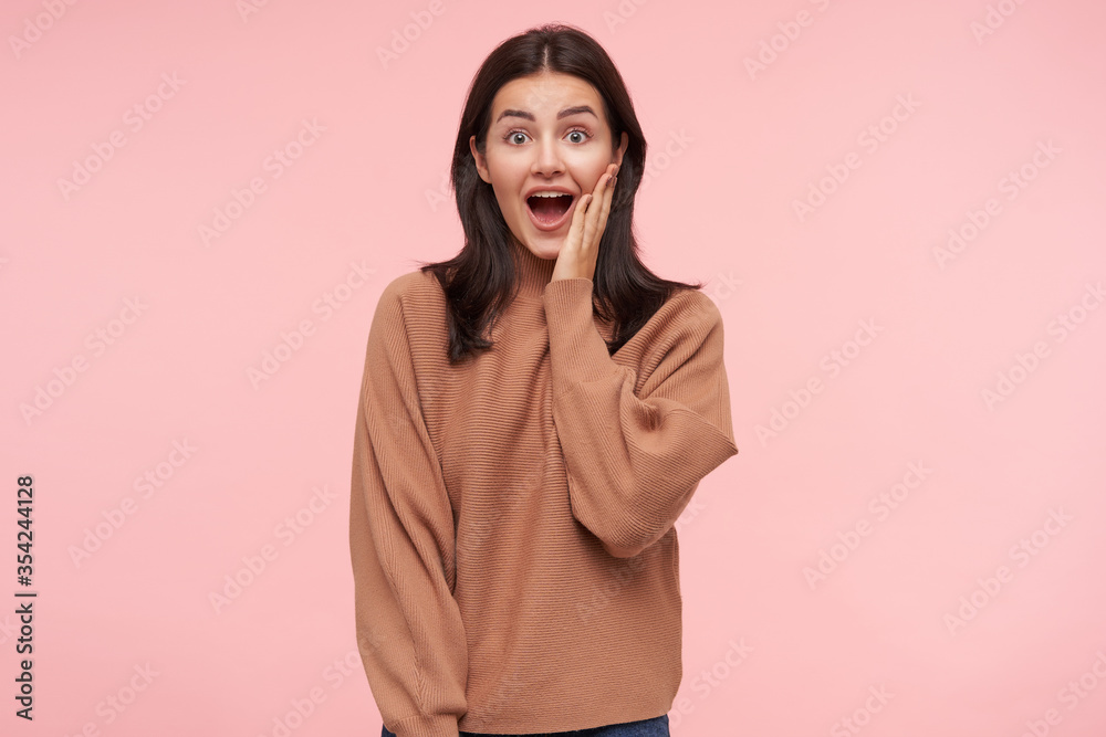 Bemused young lovely brunette female with casual hairstyle keeping her mouth wide opened while looking dazedly at camera, raising hand to her face while posing over pink background Bemused young lovely brunette female with casual hairstyle keeping her mouth wide opened while looking dazedly at camera, raising hand to her face while posing over pink background