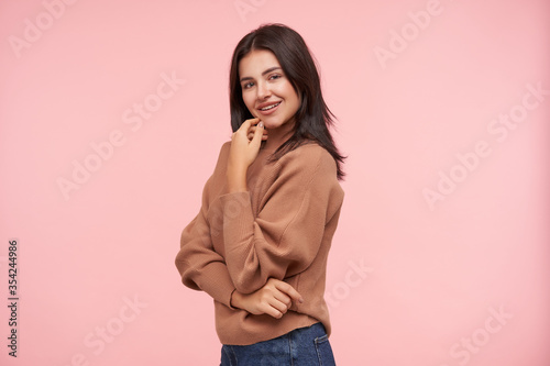 Positive young attractive brunette female with casual hairstyle touching gently her face with raised hand and smiling pleasanty at camera, isolated over pink background © timtimphoto