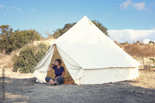 Staycations - local camping outdoor vacation. Woman drinking tea near big glamping tent with cozy interior. Luxury travel accomodation into the forest. Hyper-local travel  night camping out concept