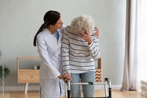 Attentive nurse giving psychological physical support to unhappy aged disabled crying woman, patient holding walking frame suffers from disease, understanding overcome together, rehabilitation concept
