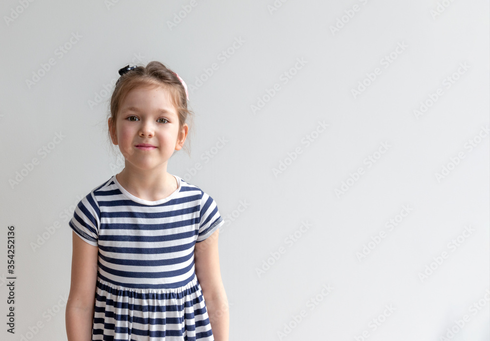 Charming little girl stands in a striped dress standing against a gray wall. International Children's Day.