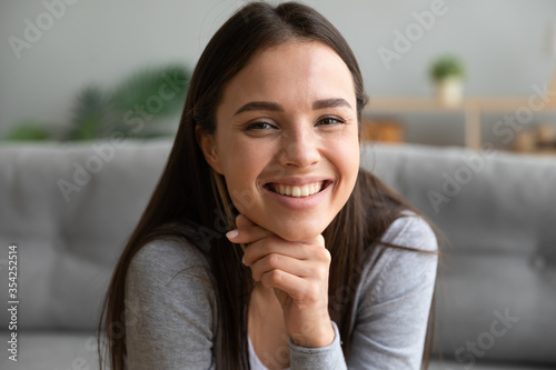 Beautiful brown-eyed woman sit on couch puts hand on chin smiles looks at camera, millennial generation representative, female has white teeth perfect skin natural beauty, optimism good mood concept