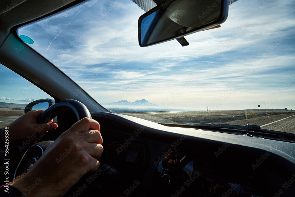Fototapeta premium Windscreen view of nature, road, sky and hand of man on the steering wheel in a spring, summer or autumn day. The concept of a single trip