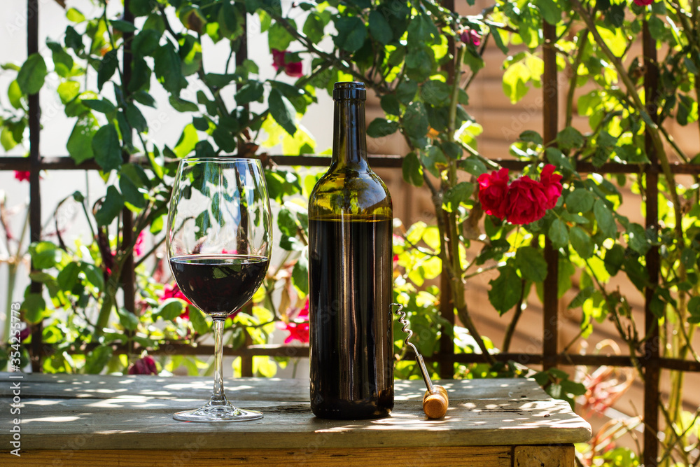 Botella de vino tinto con una copa sobre una mesa de madera rústica y un fondo de naturaleza con flores. Vista de frente