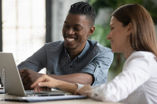 Happy diverse colleagues working on online project together, using laptop, looking at screen, discussing strategy, sharing creative ideas, employees having fun during break, laughing at joke