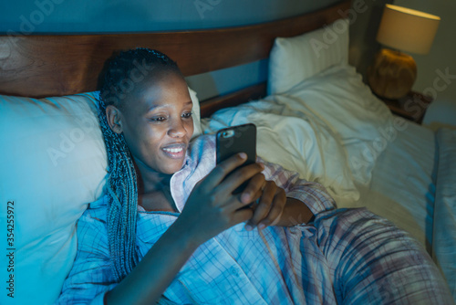 happy African American millennial woman as social media addict - night lifestyle portrait of young beautiful and cool black girl texting or online dating on mobile phone in bed