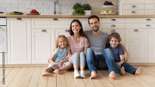 Portrait of young happy family with small children relax on wooden floor in new design kitchen, smiling Caucasian parents rest enjoy leisure weekend with little kids in rent renovated home or studio