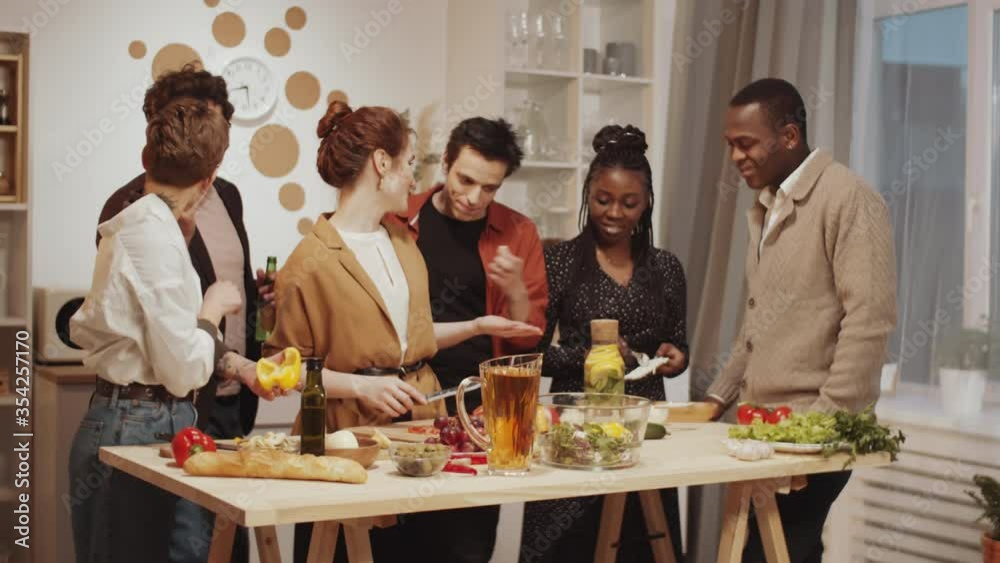 Company of young diverse friends smiling and joking while getting ready for home dinner together: cheerful women cooking food while their boyfriends having beer and stealing some vegetables Company of young diverse friends smiling and joking while getting ready for home dinner together: cheerful women cooking food while their boyfriends having beer and stealing some vegetables