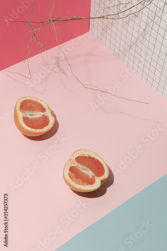 Branch and halves of grapefruit on a light pink blue surface in front of geometric perspective background. Direct hard light