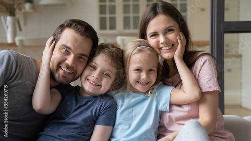 Portrait of happy young family with little children sit on couch hug cuddle look at camera posing, smiling parents have fun rest at home with small preschooler kids brother and sister enjoy weekend