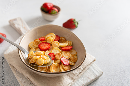 Pancake cereal, mini pancakes in a bowl with strawberries on white background. Trendy food during quarantine and lockdown. Side view, copy space. Menu, recipe, banner. Kid's breakfast.