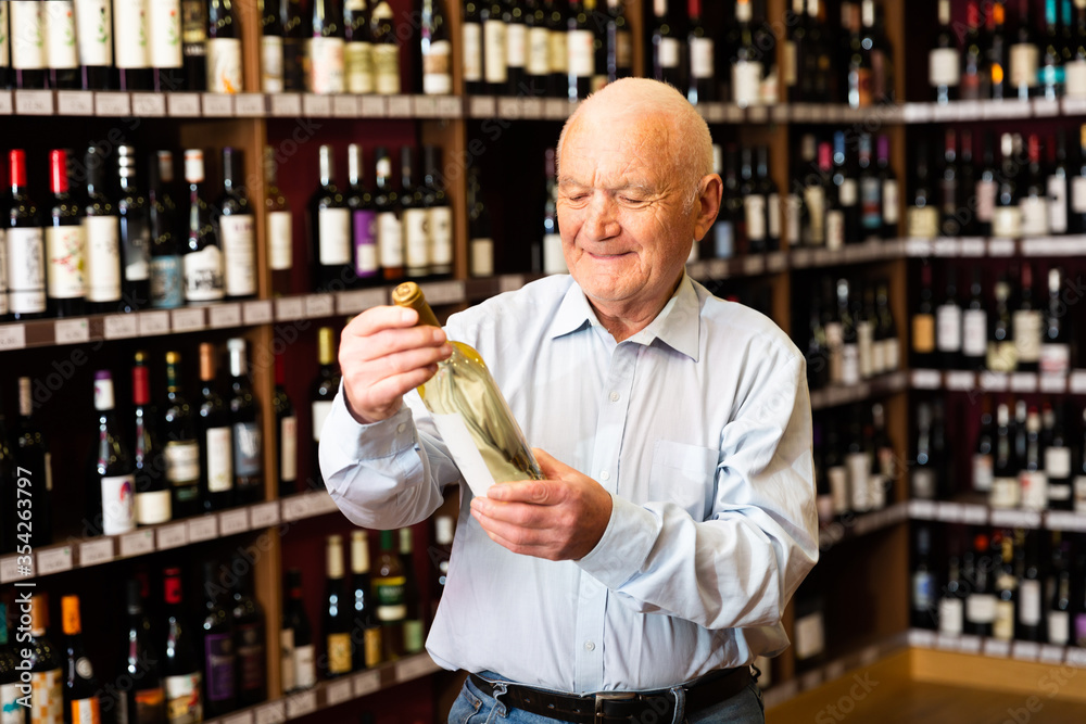 Confident grey-haired elderly man choosing white wine in modern wineshop