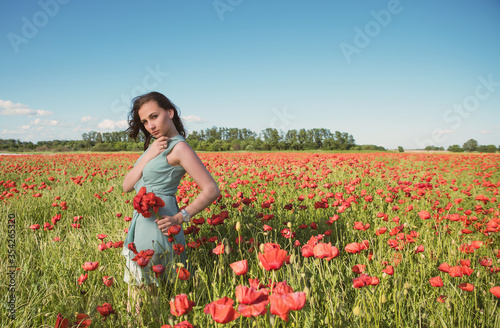 A beautiful, slender girl in a short turquoise dress against the background of a poppy field in the rays of the setting sun