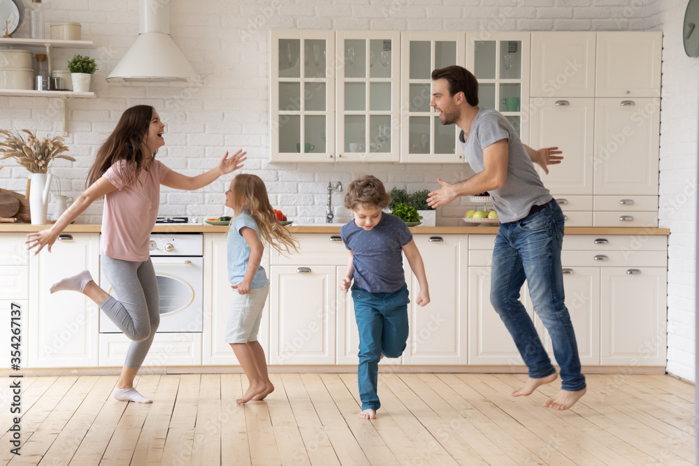 Naklejka premium Overjoyed young family with little children dancing enjoying leisure weekend in kitchen, happy parents have fun feel playful engaged in funny activity with small kids at home, entertainment concept