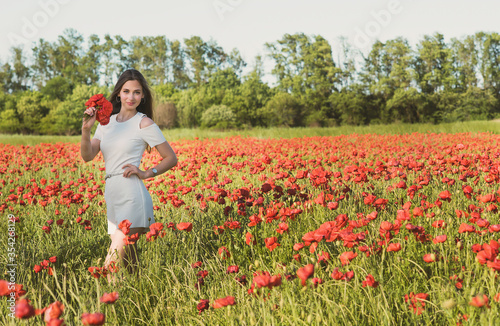 A dark, stylish, slender girl poses against the background of a poppy field with her hair flowing in the wind in the warm rays of the summer sun