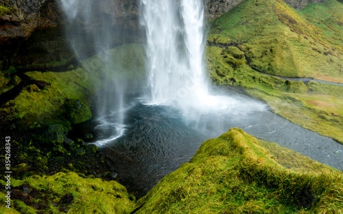 view of the popular magical Seljalandsfoss waterfall cascading into a small pond surrounded by a footpath trough green grass, nearby Storidalur, iceland