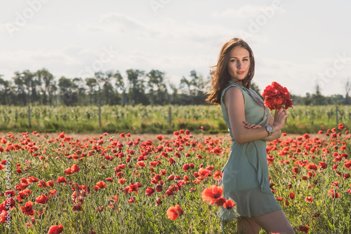 A beautiful, slender girl in a short turquoise dress against the background of a poppy field in the rays of the setting sun