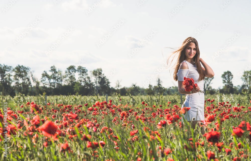 A dark, stylish, slender girl poses against the background of a poppy field with her hair flowing in the wind in the warm rays of the summer sun