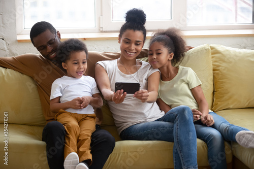 African parents and little adorable kids sitting on sofa in cozy modern living room having fun make selfie using smart phone. Chatting with relatives via videocall, modern tech family weekend concept