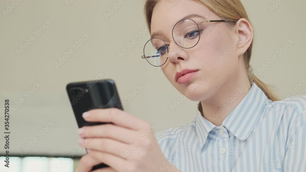 A friendly young woman architect wearing glasses is using her smartphone while sitting in the office A friendly young woman architect wearing glasses is using her smartphone while sitting in the office