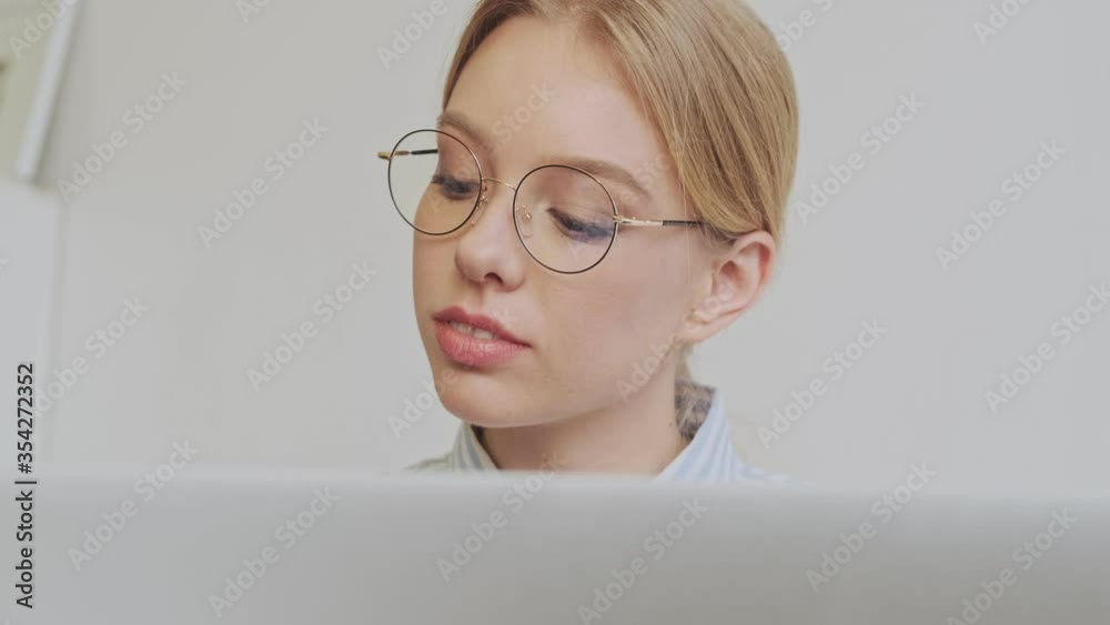 A close-up view of a beautiful young blonde woman architect wearing glasses is working with her laptop sitting in the modern office A close-up view of a beautiful young blonde woman architect wearing glasses is working with her laptop sitting in the modern office