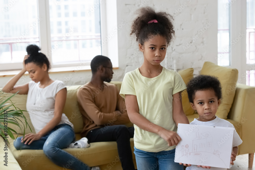 Unhappy little African siblings holding drawing where happy full family and house looks at camera. Psychological Effects of divorce on kids, parents separation break up, dysfunctional family concept Unhappy little African siblings holding drawing where happy full family and house looks at camera. Psychological Effects of divorce on kids, parents separation break up, dysfunctional family concept