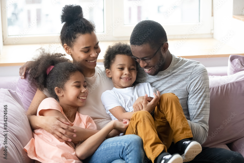 African ethnicity parents pre-teen daughter little son having fun sitting on couch tickling each other laughing enjoy priceless time together. Affection and connection, family ties and bond concept African ethnicity parents pre-teen daughter little son having fun sitting on couch tickling each other laughing enjoy priceless time together. Affection and connection, family ties and bond concept