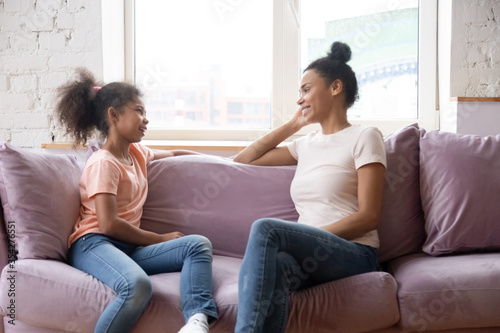 African family spend time at home mother sitting on couch with pre-teen daughter women talking smiling enjoy pleasant honest conversation. Communication and understating between two generation concept