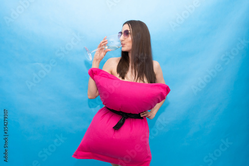 happy woman in pink pillow dress and sunglasses drinks a glass of champagne on a blue background for the crazy Pillow Challenge due to being at home in isolation. Fashionable girl. Pillow call due to photo