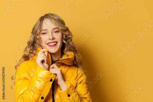 Fotografie Young smiling woman in winter jacket on bright yellow background, close up portr