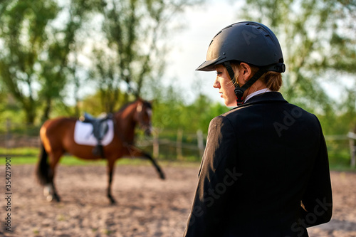 A horse rider girl stands on a farm wearing a helmet on his head. In the background stands a horse raised his leg.
