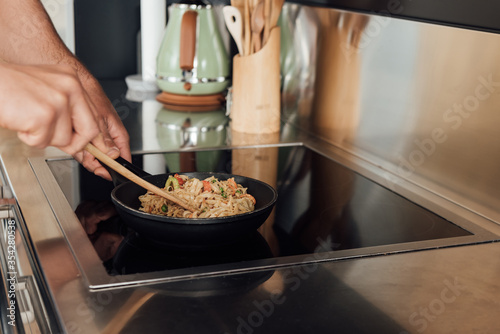 Cropped view of man holding wooden spatula while preparing noodles in frying pan in kitchen
