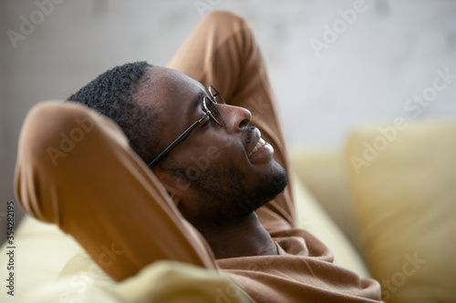 Close up image peaceful young african ethnicity man in glasses closed eyes put hands behind head resting leaned on couch at modern home. Enjoy fresh air, reduces fatigue, no stress lifestyle concept