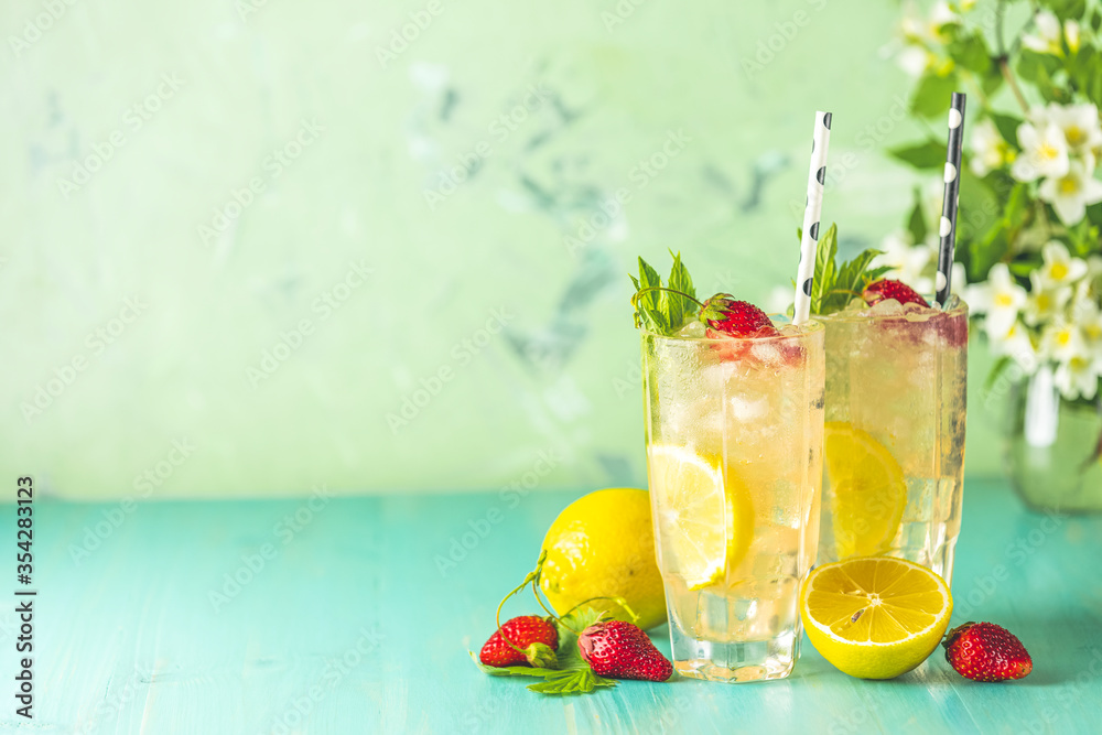 Two glasses of cold icy refreshing drink with lemon and strawberry served with bar tools on green wooden table with white blooming flowers. Fresh cocktail drinks with ice fruit and herb decoration. Two glasses of cold icy refreshing drink with lemon and strawberry served with bar tools on green wooden table with white blooming flowers. Fresh cocktail drinks with ice fruit and herb decoration.